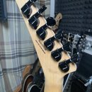 Detail hlavy elektrické kytary z přírodního světlého dřeva s šesti černými ladičkami. Na zadní straně hlavy je nápis ISJ2303199 a CRAFTED IN INDONESI…