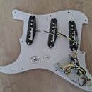 Bílá pickguard s třemi instalovanými snímači typu single-coil. Na povrchu je kulatý razítkový nápis QC PSE INSPECTION a malá papírová nálepka s označ…
