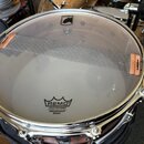 Fotografie zobrazuje snare buben s průhlednou blánou. Na bláně je černý nápis REMO WEATHERKING AMBASSADOR SNARE a v horní části logo MAPEX s ikonou k…