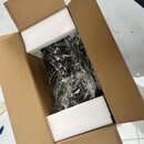 V hnědé papírové krabici s bílými polystyrenovými vložkami se nachází černý hudební přístroj. Nástroj je zabalen v průhledné plastové fólii, která za…