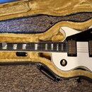 Bílá elektrická kytara s tvarem single-cut v černém kufru s žlutým vnitřkem. Na hlavě je logo Epiphone a nápis "Elite". Kytara má dva humbuckery, zla…