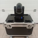 Černý LED reflektor typu moving head umístěný na černé transportní bedně s hliníkovými hranami a koly. Na zadní straně reflektoru jsou viditelné nápi…