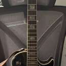 Černá elektrická kytara s tvarem typu Les Paul a zlatým hardwarem. Na hlavě je viditelné logo "Epiphone". Hmatník má vložky z perlece nebo podobného…