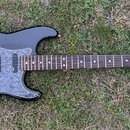 Černá elektrická kytara s šedým mramorovaným pickguardem a hnědým hmatníkem. Na hlavě je viditelné logo Fender a nápis STRATOCASTER, níže je drobný n…
