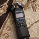 Černý digitální záznamový přístroj značky zoom. Na přední straně je displej, ovládací tlačítka a v dolní části nápis H1essential Handy Recorder. V ho…