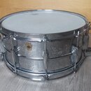 Kovový snare drum s chromovaným povrchem a bílým bubenovou skins. Na korpusu je viditelné zlaté logo ve tvaru štítu, ale text uvnitř něj není čitelný…