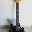 Černá elektrická basová kytara s přírodním dřevěným hmatníkem a hlavou. Na hlavě je nápis Fender MUSTANG BASS. Nástroj má černý plech ochranný kryt (…