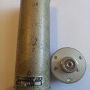 Cylindrický mikrofon v matné zlatavé barvě s viditelným opotřebením povrchu a škrábancem. Na spodní části je štítek s nápisy: G.NEUMANN & CO. GMBH i.…