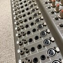 Detailní pohled na míchací pult s kovovým šedým povrchem. Jsou vidět řady konektorů, včetně vstupů MIC (XLR), LINE IN a INSERT, s popisky D OUT, TAPE…