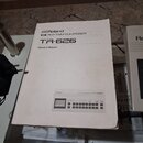 Na fotografii je bílý printed manuál s nápisy Roland MIDI RHYTHM COMPOSER TR-626 Owner's Manual a obrázkem přístroje. Vedle manuálu je umístěn bílý e…