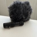 Černý mikrofon s nasazenou šedou chlupatou ochranou proti větru. Na ochraně je viditelné logo RØDE a na těle mikrofonu nápisy Videomic Me MADE IN AUS…