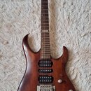 Elektrická kytara s hnědým lakovaným tělem a tmavým hnedým hmatníkem. Na hlavě je bílý nápis Cort. Nástroj má tři humbuckery, tremolo most a dvě ovlá…