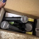 V kartonové krabici se nacházejí tři černé dynamické mikrofony s kovovými mřížkami. Na dvou z nich je viditelný nápis SHURE SM58. Mikrofony jsou ulož…