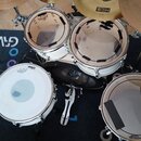 Bílá bubnová souprava s několika činely. Na blachách jsou viditelné loga REMO a EVANS. Jeden z činelů má nápis paiste pst 5, jiný nápis HCS.