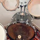 Černá bicí souprava s kovovými obručmi a mechanismy. Na dvou tom-tamech i na basovém bubnu je vidět nápis SONOR. Basový buben stojí na červeném vzoro…