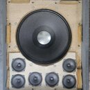 Šedý dřevěný rozměrný box s vnitřním polstrováním, ve kterém je osazen jeden velký černý subwoofer a šest menších černých reproduktorů. V horní části…