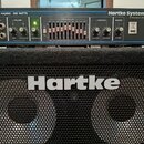 Kombinovaná fotografie basového zesilovače a reproduktorové skříně. Zesilovač má modré tělo s nápisy MODEL HA3500 350 WATTS a Hartke Systems Transien…
