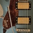 Detail elektrické kytary v modré metalické barvě s hnědo-červeným vzorovaným pickguardem, na kterém je bílý nápis GRETSCH. Nástroj má dva humbuckery…
