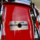 Detail červeného bubnu s chromovaným obručem a lugy. Na korpusu je kovový štítek s nápisy YAMAHA, MODEL S0098, SER. NO. NY6970 a MADE IN JAPAN. Povrc…