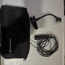Černý mikrofon s ohebným hrdlem a připojeným kabelem. Vedle něj je černý přepravní pouzdro s bílým nápisem beyerdynamic))). Vše leží na stříbrném pov…