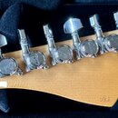 Zadní strana světle hnědé dřevěné hlavy kytary s šesti chromovanými ladičkami. Na zadní straně každé ladičky je vyryto logo "Fender". V dolní části h…
