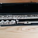 Stříbrná flétna rozložená v černém kufru. Nástroj je tvořen několika částmi s kovovými klapky a mechanismy. Na povrchu nejsou viditelné žádné značky…