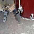 Detail červeného bubnu s kovovým kováním a dvěma černými držáky v podobě válců. Vedle bubnu je vidět část basového pedálu s černým šlapadlem a kovovo…