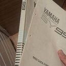 Dvě bílé tištěné příručky s nápisy. Na přední straně viditelného manuálu je logo YAMAHA, text MUSIC SYNTHESIZER SY99 a MIDI DATA FORMAT. Na hřbetu dr…