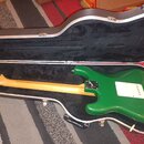Zelená elektrická kytara v černém tvrdém kufru. Na hmatníku je vidět bílý pickguard a na hlavě kytary nápis Fender. Povrch laku je hladký a bez vidit…