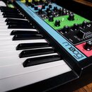 Klávesový syntetizátor s černým šasi a bílo-černými klávesami. Na modrém panelu je viditelné logo moog a nápisy SUSTAIN, VOL VOLUME. Ovládací panel o…