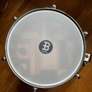 Fotografie zachycuje bílou bubenovou membránu s kovovým obručem. Uprostřed je černé kulaté logo s bílým symbolem „B“ a nápisem MEINL. Membrána je v č…