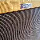 Detail kytarového komba s žlutým texturovaným povrchem a tmavou mřížkou reproduktoru. Na horní části je umístěno stříbrné logo s nápisem Fender. V ho…