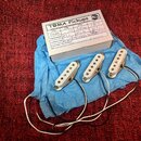 Tři bílé jednocitové snímače s kovovými šrouby, položené na modrém papíře. Vedle nich je kartička s nápisem TOMA Pickups a tabulkou, ve které jsou ru…