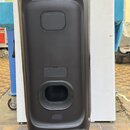 Černý party reproduktor na kolečkách s nápisem PARTYBOX v horní části. Povrch je matný, v zadní části jsou vidět dva obdélníkové otvory a jeden kulat…