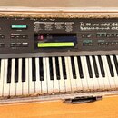 Černý digitální syntetizátor Yamaha DX7 II-D v hliníkovém transportním kufru. Na ovládacím panelu je LCD displej s textem "Voice: Internal Inf 5 Full…