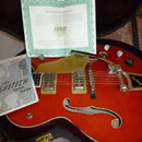 Červená polodublová elektrická kytara značky Gretsch v otevřeném kufru. Na těle jsou dva snímače, Bigsby vibrato a nápis Gretsch na zlatém krytu. K n…