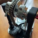Stříbrný kovový mechanismus s řetězem, který je umístěn na černém stojanu s bílou základnou. Na kovovém prvku je nápis „INFINITE ADJUSTABLE CAM“. Na…