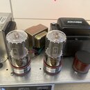 Detail vnitřku elektronického zařízení s dvěma elektronkami, transformátory a hnědým válcovým kondenzátorem. Na elektronkách jsou viditelné nápisy „6…