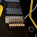 Detail elektrické kytary v černé barvě s žlutými pruhy. Nástroj má dva černá snímače s nápisem EMG a chromovaný tremolo systém. Na těle jsou vidět dv…