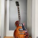 Elektrická kytara s tvarem singlecut a povrchovou úpravou v hnědém sunburstu. Na hlavě je viditelné logo Gibson. Kytara má dva humbuckery, čtyři otoč…