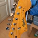 Zadní strana hlavy kytary v žlutém lakovaném provedení s viditnou strukturou dřeva. Na zadní straně je černý nápis Squier JAZZMASTER by Fender. Jsou…