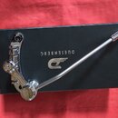 Chromovaný kytarový tremolo systém položený na černé krabici. Na krabici je bílý nápis DUESENBERG a logo ve tvaru stylizovaného písmene D s křídly. P…