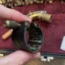 Černý kožený ligaturek na saxofon s kovovým šroubem. Na vnitřní straně je zlatým nápisem vyrazen text: L12SR SAX ALTO SUPER REVELATION. V pozadí je v…