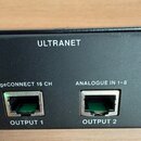Detailní záběr na zadní stranu černého elektronického zařízení. Jsou zde vidět dva ethernetové porty s nápisy OUTPUT 1 a OUTPUT 2. Nad porty jsou bíl…