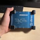 Modrá DI box s černými gumovými nárožníky. Na horní straně je bílý nápis SAMSON a svisle orientovaný nápis S-direct. Uprostřed je schéma zapojení s t…