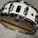 Bílá buben (snare), s chromovanými obručmi a černými ladicími mechanismy. Na spodní straně je viditelné logo MAREX v čtvercovém rámečku. Povrch nástr…