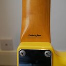 Detail zadní strany krku a spoje s tělem žluté kytary. Na dřevěném krku je černý nápis Crafted in Japan. V dolní části je vidět kovový kryt připevněn…