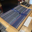 Modrý mixovací pult s velkým množstvím potenciometrů a posuvníků. Uprostřed je viditelné logo TASCAM. Pult je umístěn na dřevěném stole vedle počítač…