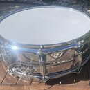 Kovový snare drum s chromovaným povrchem a bílou blánou. Nástroj je umístěn na dřevěném stole venku. Na těle bubnu nejsou viditelné žádné značky ani…