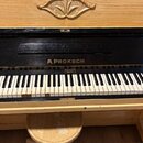 Dřevěný piano s černým lakovaným povrchem v oblasti klaviatury. Nad klávesami je zlatý nápis A.PROKSCH a pod ním malý, nečitelný štítek. Na horní dře…